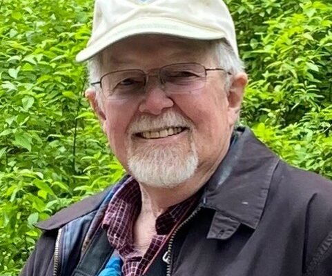 A photo of Riley smiling with a baseball cap and green leafy background.